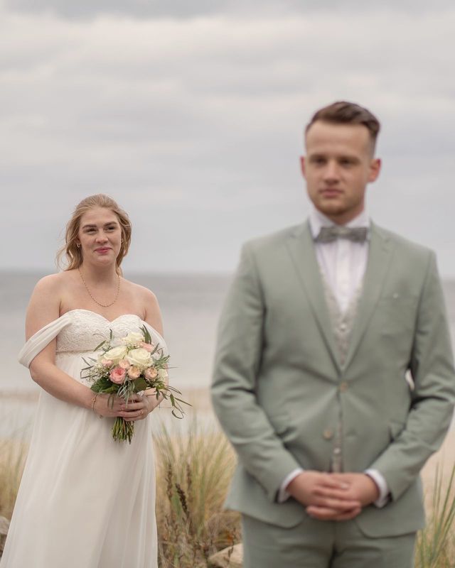 Braut mit Strauß in den Dünen am Meer, vorn unscharf der Bräutigam.