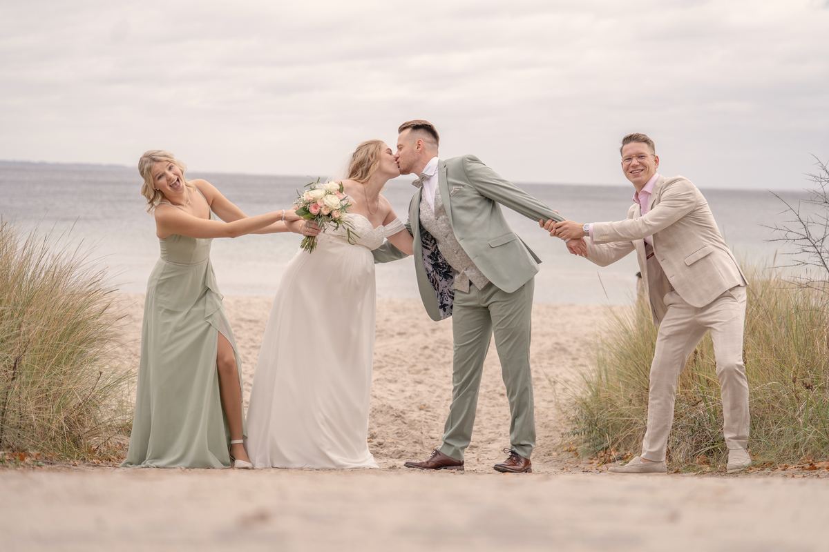 Zwei Freunde ziehen das küssende Brautpaar am Ostseestrand an den Händen.