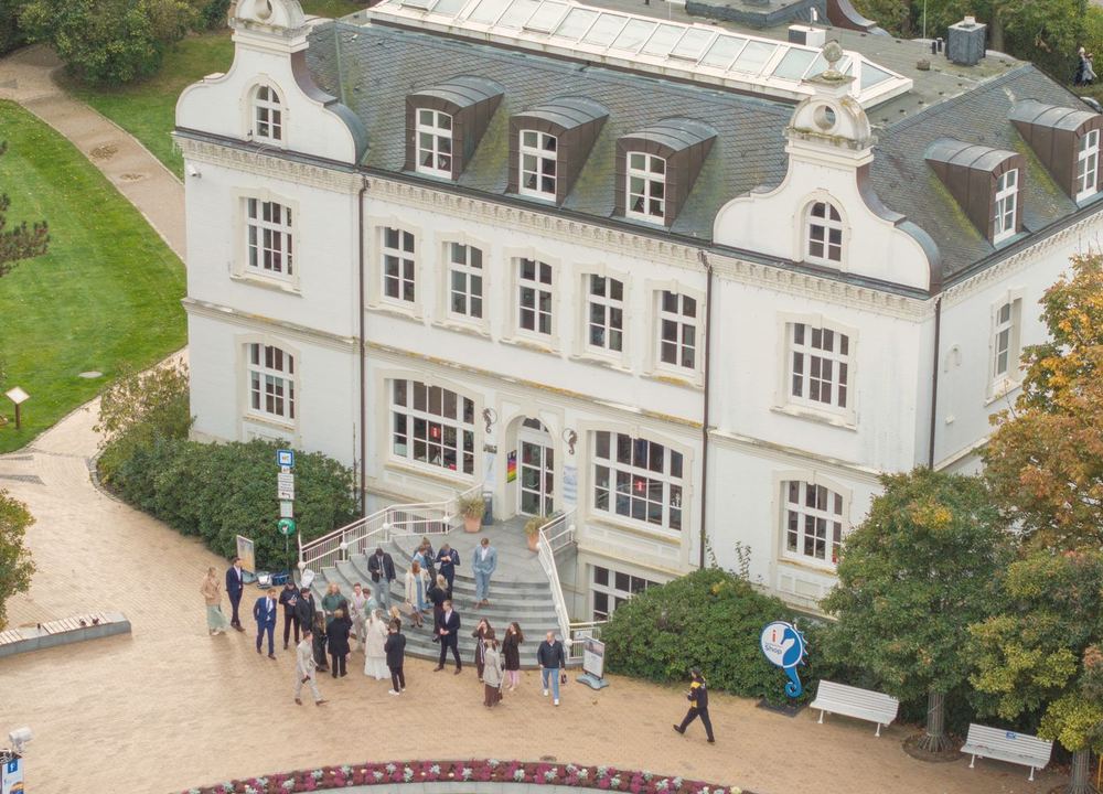 Luftaufnahme einer weißen Villa, auf der Treppe davor stehen die Hochzeitsgäste.